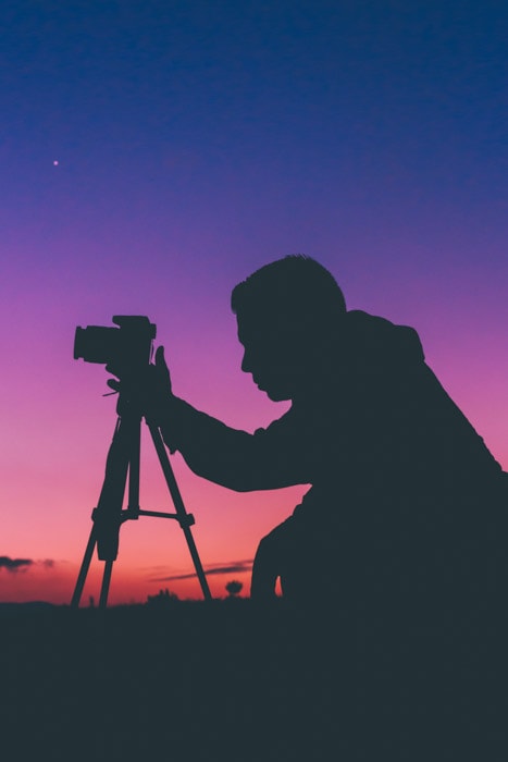 Night time silhouette of a photographer kneeling at his camera on a tripod with a brilliantly colored sky behind him